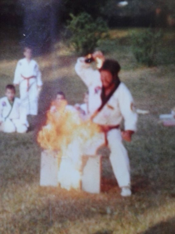 A grainy outdoor photo: Master Birch, mid-strike, breaks a stack of boards that bursts into flame while students in uniforms watch from the grass.